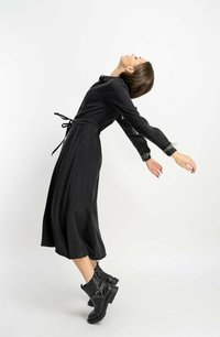 Czarna sukienka z długim rękawem z talią ściągniętą i lejącą się spódnicą; ma szczegółowe mankiety i jest noszona z masywnymi, czarnymi botkami za kostkę.