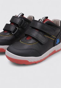 Baskets noires en cuir et tissu texturé. Elles présentent deux bandes Velcro, des accents en maille rouge, des coutures jaunes et une semelle en caoutchouc avec des détails rouges.