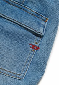 Tessuto di denim blu chiaro con una texture vissuta; presenta cuciture arancioni e un logo ricamato rosso vicino alla tasca.