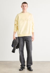 Sweatshirt amarelo com logótipo "HUGO" em relevo, combinado com jeans cinzentos escuros. O modelo segura um casaco cinzento. Design simples e descontraído.