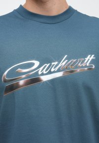 Camiseta de algodón color verde azulado con un logo de Carhartt en plata metálica, acabado brillante y detalles reflectantes sutiles. Manga corta y cuello redondo.