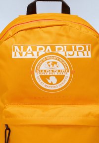 Mochila de tela amarilla con un logotipo frontal que presenta un globo terráqueo y la Antártida. Incluye un asa superior y un zipper rojo en la parte superior.