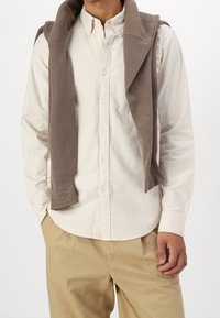 Beige skjorte med knapper og krave, kombineret med en brun sweater, der hænger over skuldrene, og lyse khaki bukser.