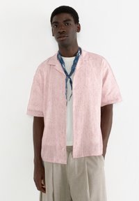 Cămașă roz cu mânecă scurtă și nasturi, cu un model abstract alb, purtată peste un tricou alb, asortată cu pantaloni bej deschis și o eșarfă albastră.