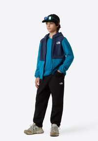 Blå och marinblå zip-up-hoodie över en teal t-shirt, kombinerad med svarta byxor och gråa vandringsskor. Outfiten har logodetaljer.