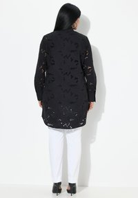Tunic nera ricamata con motivi floreali a taglio e maniche lunghe, abbinata a pantaloni bianchi. Scarpe nere con tacco completano l'outfit.