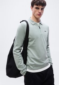 Νεαρός άνδρας που φοράει ανοιχτό πράσινο πουλόβερ με φερμουάρ και μαύρο παντελόνι, κρατώντας μια μεγάλη μαύρη τσάντα ώμου, στέκεται μπροστά σε ένα απλό φόντο.