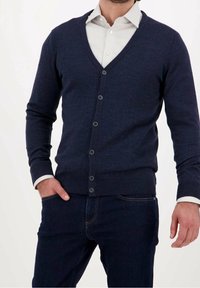 Cárdigan azul marino de tela de punto con cuello en V, seis botones, puños y ribete acanalados, superpuesto sobre una camisa blanca. Textura distintiva.