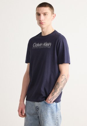 T-shirt in cotone blu navy con logo bianco "Calvin Klein" sul petto, maniche corte e vestibilità rilassata, abbinato a pantaloni corti in denim blu chiaro.
