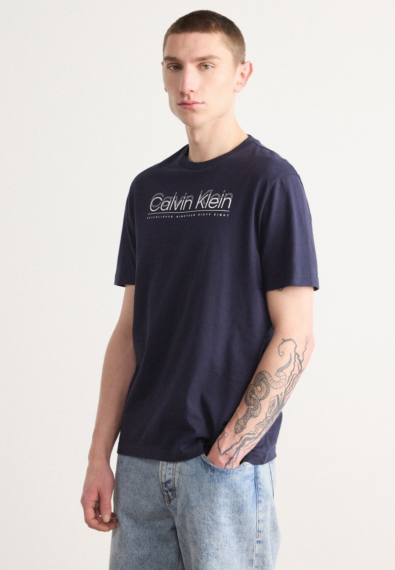 Navyblauwe katoenen t-shirt met een witte "Calvin Klein" logo op de borst, korte mouwen en een relaxte pasvorm, gecombineerd met lichtblauwe denim shorts.
