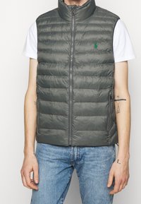 Grå vadderad väst med hög krage, dragkedja fram, sidofickor och liten broderad logotyp på vänster bröst, bärs över en vit skjorta.