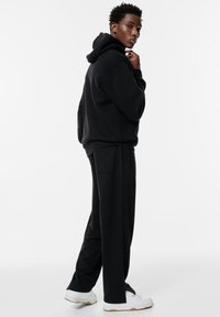 Schwarzer Hoodie mit lockerer Passform und Kapuze, kombiniert mit passenden schwarzen Jogginghosen. Weiße Sneakers vervollständigen das Outfit. Glatte Stofftextur.