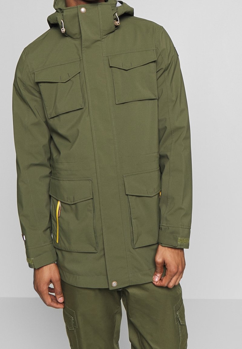 Homme portant une veste à capuche vert olive avec quatre poches à rabat et un pantalon cargo assorti sur fond gris uni.