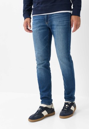 Mann trägt blaue Slim-Fit-Jeans, marineblaue und weiße Sneakers, steht mit entspannten Armen an den Seiten vor weißem Hintergrund.