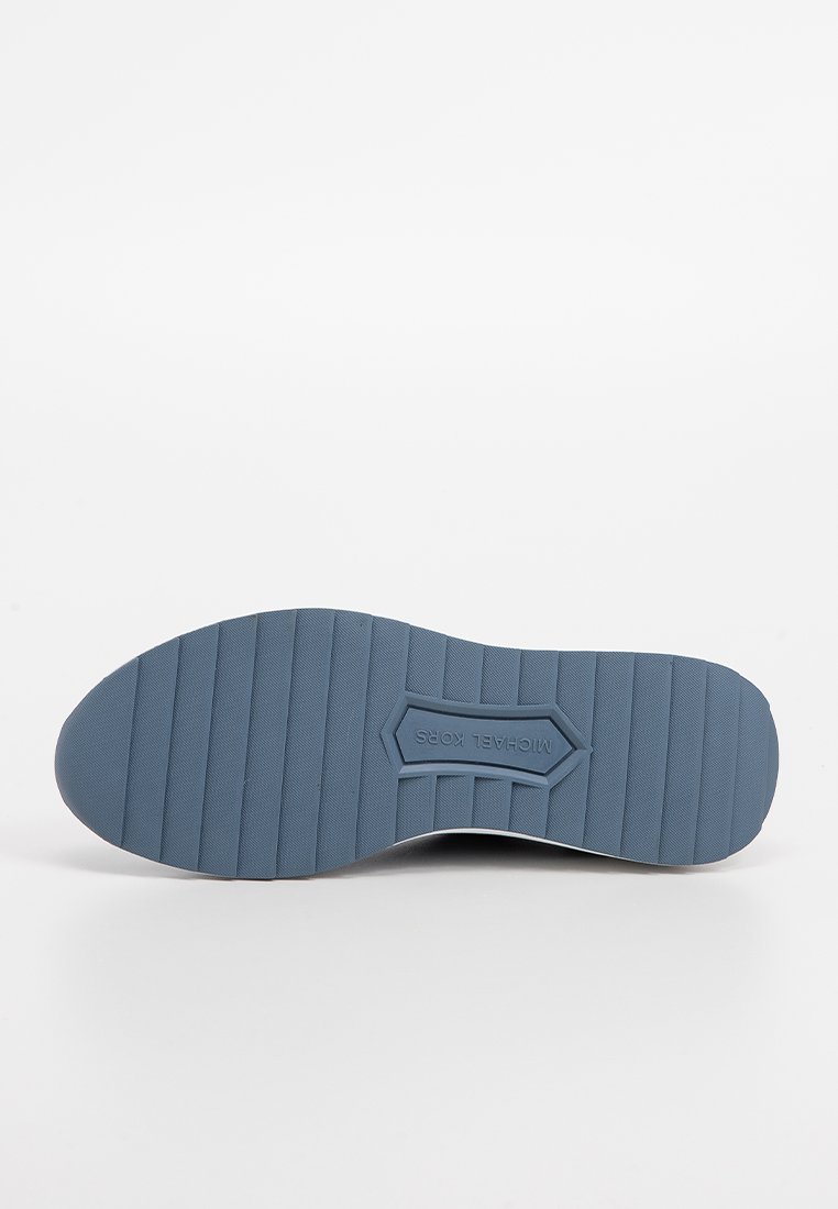 Semelle de chaussure en caoutchouc grise avec des rainures horizontales pour une meilleure traction, présentant un logo hexagonal au centre. Texture lisse et forme arrondie.