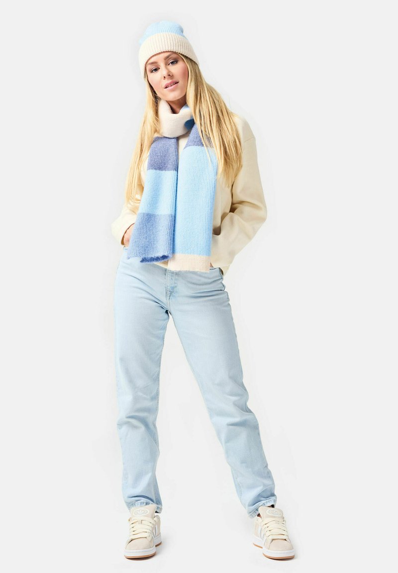 Hellblau gestrickte Mütze mit Bommel, gestreifter hellblauer und cremfarbener Schal, cremefarbener Pullover, hellblaue Jeans und weiße Sneakers.