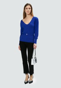 Suéter azul de cuello en V con textura acanalada, cintura ceñida, combinado con pantalones acampanados negros con borde de encaje y tacones negros con acentos plateados.