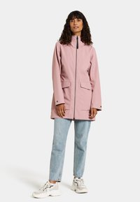 Lichtroze waterdichte regenjas met een hoge kraag, voorzijde met rits, twee grote zakken en een relaxte pasvorm, gecombineerd met blauwe jeans en sneakers.