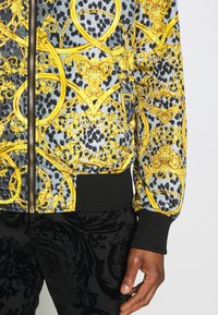 Veste noire et grise à motifs avec des dessins baroques jaunes ornés et des poignets côtelés noirs, portée avec un pantalon noir à texture florale. Main visible.