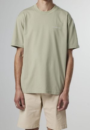 T-shirt en coton vert clair à manches courtes, col rond, avec un texte brodé sur la poitrine gauche. Associé à un short beige.