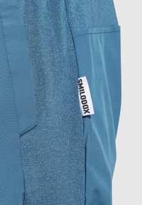 Pantalones deportivos azules hechos de un material suave, con un patrón texturizado, un bolsillo lateral y una etiqueta con "SMILODOX" en colores contrastantes.