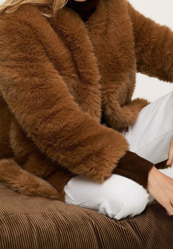 Brązowy płaszcz z sztucznego futra o puszystej fakturze, szerokim kołnierzu i długich rękawach. Połączony z białymi spodniami, na tle pr�ążkowanego brązowego tła.