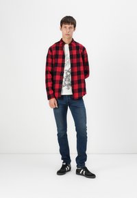 Červená a čierna károvaná flanelová košeľa, biela grafická tričko pod ňou, modré džínsy a čierne tenisky s bielymi pruhmi. Neformálny outfit.