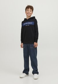 Sudadera negra con gráfico de rayas azules y diseño de texto, combinada con pantalones vaqueros de mezclilla azul holgados y zapatillas blancas. Tejido suave y corte relajado.