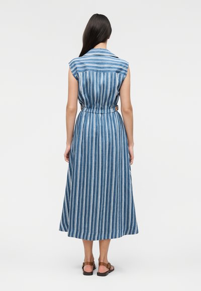 Γυναίκα με μακριά σκούρα μαλλιά, φορώντας ένα μίντι φόρεμα χωρίς μανίκια με μπλε και λευκές κάθετες ρίγες και καφέ επίπεδα σανδάλια, στέκεται γυρισμένη προς τα πίσω.