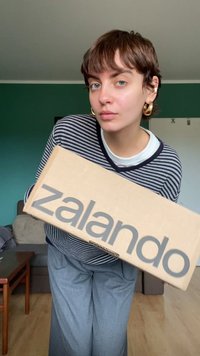 Zalando pappkarp, mida hoiab inimene, millel on lihtne beež värv ja suur hall logo tekst. Inimene kannab triibulist särk ja pükse.