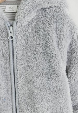 Harmaa fleece-takkia, jossa on pehmeä ja teksturoitu pinta. Varustettu etuvetoketjulla ja tarralla, joka ilmoittaa merkin ja hoito-ohjeet.
