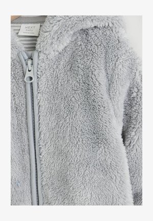 Chaqueta con capucha de felpa gris, con una superficie suave y texturizada. Cuenta con un cierre frontal de cremallera y una etiqueta que indica la marca y las instrucciones de cuidado.