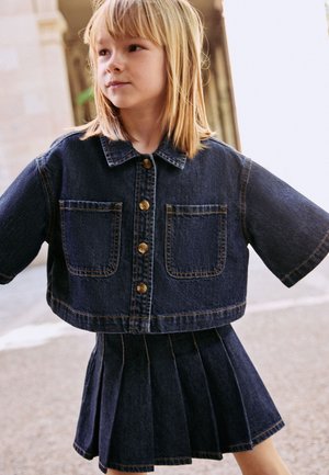 Ragazza giovane che indossa una giacca corta in denim scuro e una gonna plissettata coordinata, in piedi all'aperto con le braccia leggermente aperte.