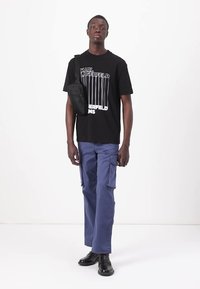 Svart bomull t-skjorte med hvite vertikale striper og tekst, parret med blå cargo bukser og svarte sko. Crossbody-veske brukt på skulderen.