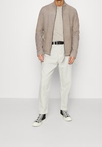 Světlá béžová semišová bunda na zip přes šedé tričko, v kombinaci s krémovými kalhotami a černými vysokými teniskami s bílými podrážkami.