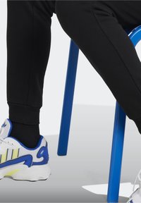 Bílé a modré sportovní boty se žlutými akcenty, zkombinované s černými zúženými kalhotami, odpočívající na modré židli.