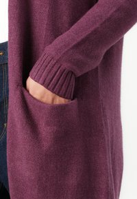 Hand in de zak van een paars gebreide trui met lange mouwen en ribgebreide manchetten, gecombineerd met donkerblauwe denim jeans tegen een witte achtergrond.