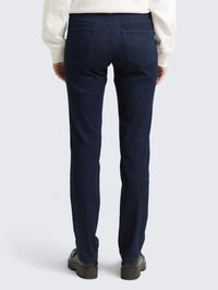 Pessoa vestindo jeans slim fit azul escuro, sapatos pretos e uma blusa de mangas longas de cor clara, de pé de costas em um fundo simples.