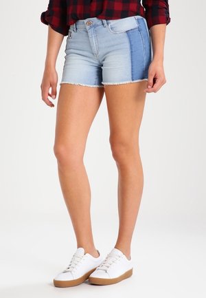 Onderlichaam van een persoon die licht- en donkerblauwe denimshorts draagt, een rood geblokt overhemd en witte sneakers met bruine zolen die op een witte vloer staan.