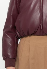 Nærbilde av en person som har på seg en maroon kunstskinn pufferjakke med ribbede mansjetter og et beige plissert skjørt.