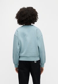 Γαλάζιο κοντό φούτερ με ριμπ μανσέτες και τελείωμα, με κλασικό στρογγυλό λαιμό και άνετη εφαρμογή. Φοράει με μαύρο παντελόνι, εμφανές ετικέτα.