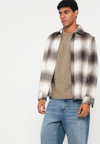 Rutete skjorte i hvit, brun og kremfarge med knapper foran, brukt over en teksturert beige genser og blå jeans.