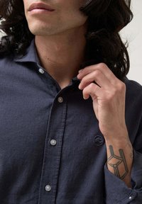 Camisa de botones de manga larga en azul oscuro, hecha de un tejido texturizado, con cuello, botonadura y un sutil detalle de logo en el pecho.