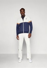 Marinblå och gräddfärgad zip-up träningsjacka med orange ränder, kombinerad med vita byxor. Har en logotyp på bröstet och detaljer på byxornas fållar.