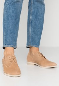 Lichtbruine suède schoenen met een gladde textuur, ronde neus en drie gaatjes voor veters, gecombineerd met blauwe denim jeans.