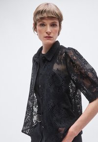 Camicia nera in pizzo con collo, caratterizzata da maniche corte e una chiusura frontale con bottoni. Il tessuto mostra motivi e trame intricati.