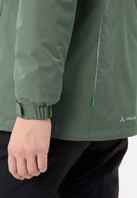 Giubbotto impermeabile verde oliva con finitura testurizzata, polsini elastici, accenti riflettenti e logo del marchio all'orlo.