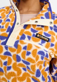 Fleece-pusero, jossa on oranssi, sininen ja kermanvärinen geometrinen kuvio. Siinä on napitus, etutasku ja Columbia-logo.