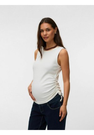 Gravid kvinde iført hvid ærmeløs top med rynker og mørke jeans, som blidt holder på sin mave, stående mod en ensfarvet lys baggrund.