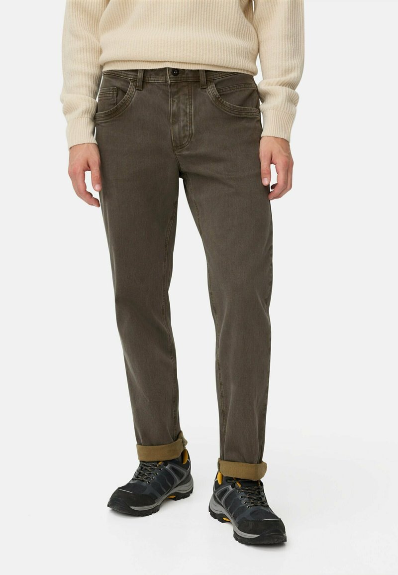 Man in opgerolde bruine broek, crèmekleurige geribde trui en zwarte wandelschoenen met gele accenten, staande tegen een witte achtergrond.